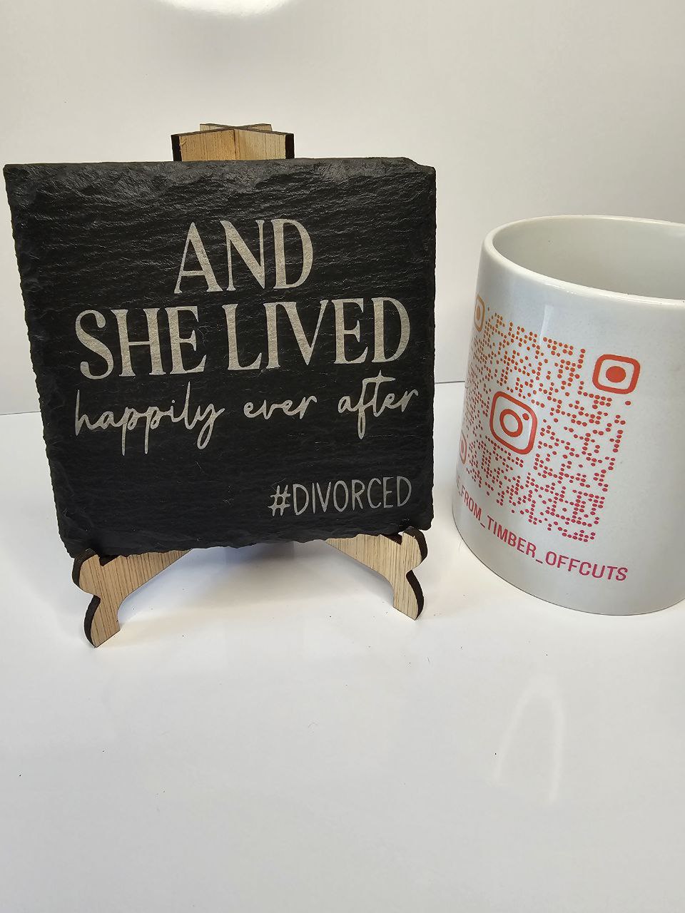 Slate coaster engraved with the phrase 'And She Lived Happily Ever After #Divorced' on a wooden stand.