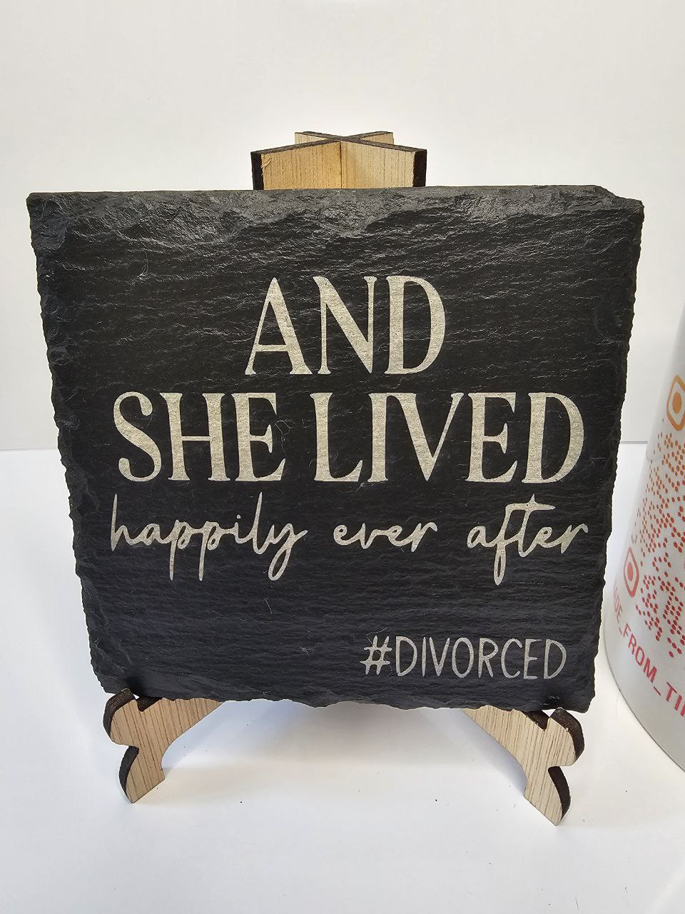 Slate coaster engraved with the phrase 'And She Lived Happily Ever After #Divorced' on a wooden stand.