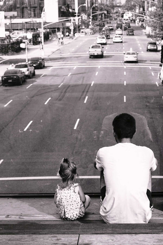 grayscale photography of man and girl sitting on staircase fronting road with passing cars