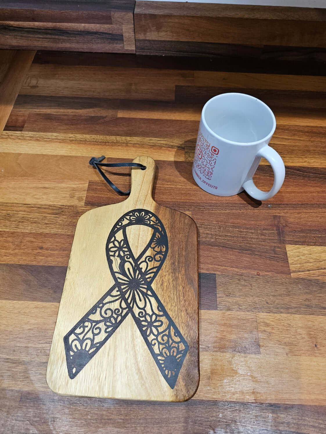 Floral Awareness Ribbon Engraved Wooden Serving Board on a wooden countertop beside a white mug.