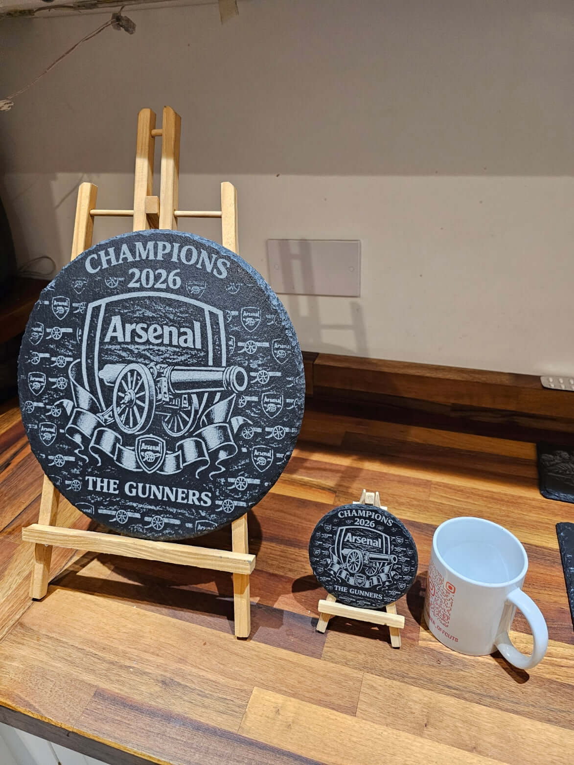 Arsenal Champions 2026 Engraved Slate Coaster displayed on an easel with a mug and a smaller coaster on the side.