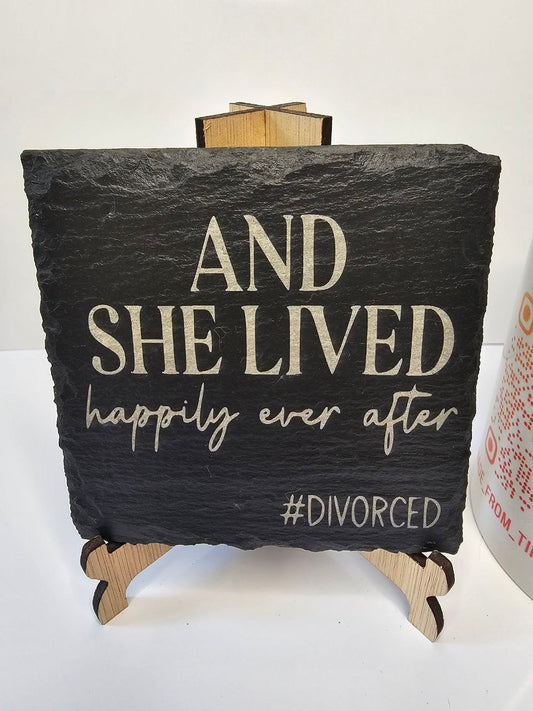 Slate coaster engraved with the phrase 'And She Lived Happily Ever After #Divorced' on a wooden stand.